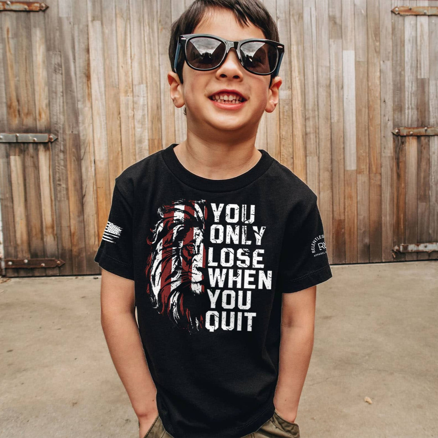 Child wearing a black t-shirt with motivational quote, standing in front of wooden shutters.